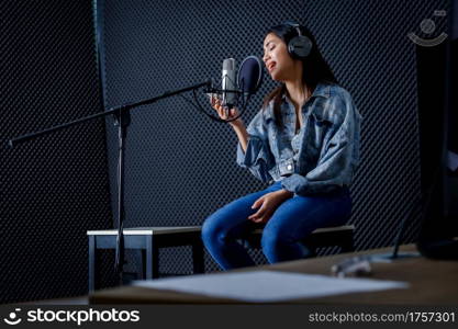 Happy cheerful pretty smiling of portrait of young Asian woman look at the smartphone vocalist Wearing Headphones recording a song front of microphone in a professional studio