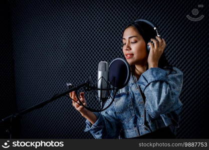 Happy cheerful pretty smiling of portrait of young Asian woman look at the smartphone vocalist Wearing Headphones recording a song front of microphone in a professional studio