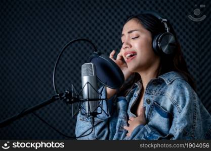 Happy cheerful pretty smiling of portrait of young Asian woman vocalist Wearing Headphones recording a song front of microphone in a professional studio