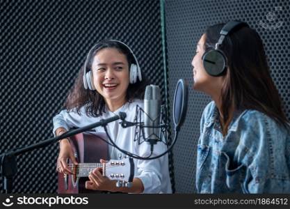Happy cheerful pretty smiling of portrait Two young Asian woman vocalist Wearing Headphones with a guitar recording a song front of microphone in a professional studio