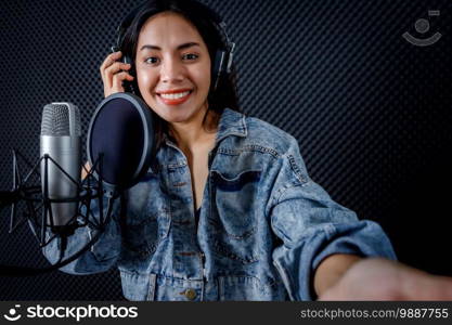 Happy cheerful pretty smiling of portrait  young Asian woman vocalist Wearing Headphones taking a selfie with smartphone while  recording a song front of microphone in a professional studio