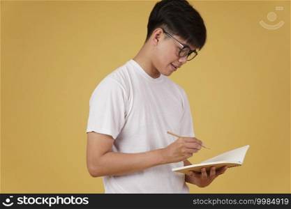 happy cheerful young asian man student dressed casually wearing eyeglasses writing note isolated on yellow studio background