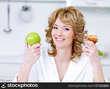 Happy cheerful young beautiful woman with green apple and sweet cake sitting in the kitchen