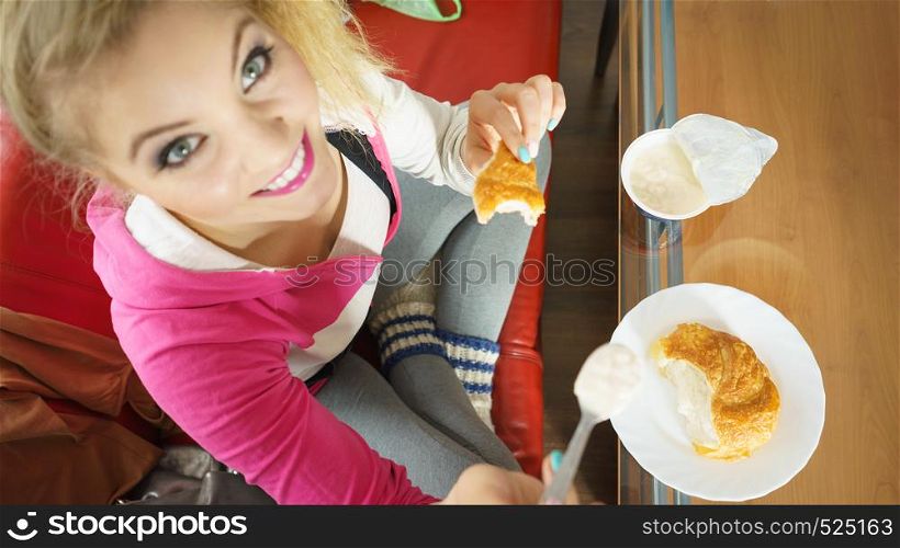 Happy cheerful young teenage woman eating croissant bun and cottage cheese yoghurt. Woman eating croissant and yoghurt