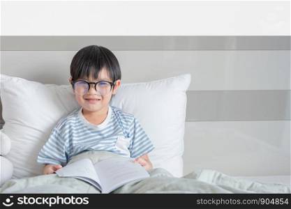 happy child little asian boy smile with holding glasses reading a books on the bedroom in room at home. family activity concept.