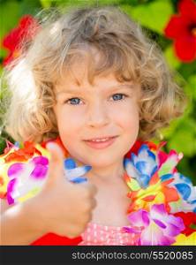 Happy child showing thumb up sign against tropical flowers background. Summer vacations concept