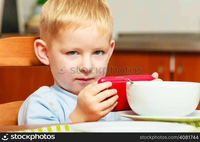 Happy childhood. Blond boy kid child eating corn flakes cereal with milk breakfast morning meal playing with mobile phone at the table. Home.