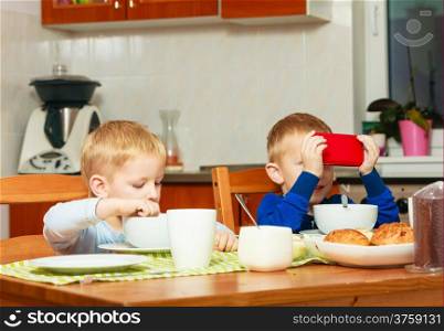 Happy childhood. Boys kids children eating corn flakes cereal with milk breakfast morning meal. Brothers playing with mobile phone at the table. Home.
