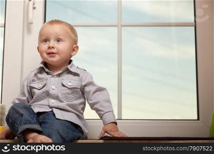 Happy childhood. Portrait of smiling little boy at home. Cute child kid in blue shirt sitting on the windowsill. Copy space.