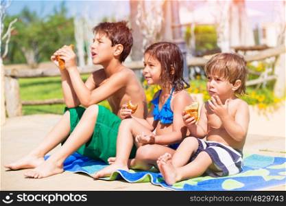 Happy children on the beach, three little best friends resting near the pool and eating croissants, enjoying active summer holidays