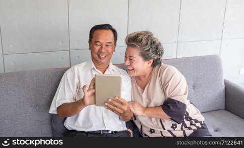 Happy couple asian senior retired using mobile tablet application technology for social network among friends community via internet digital communication while smiling and sitting on couch at their home.Asian mature couples use mobile tablets to contact online shopping
