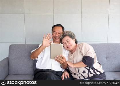 Happy couple asian senior retired using mobile tablet application technology for social network among friends community via internet digital communication while smiling and sitting on couch at their home.Asian retirees use mobile tablets to communicate with families in other cities.
