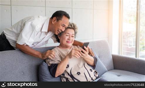 Happy couple asian senior retired using mobile tablet application technology for social network among friends community via internet digital communication while smiling and sitting on couch at their home.Asian retirees use mobile tablets to communicate with families in other cities.