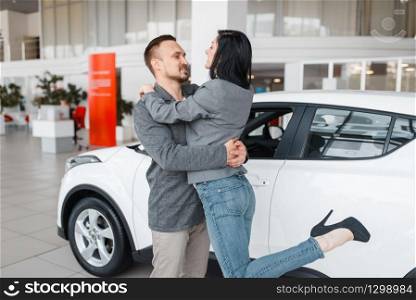 Happy couple buying new car in showroom, man and woman hugging. Male and female customers choosing vehicle in dealership, automobile sale, auto purchase