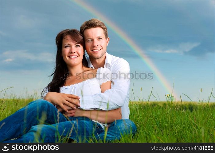 Happy couple caught in a daydream sitting on a meadow in summer; in the background a rainbow