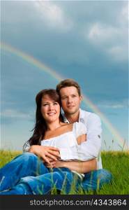 Happy couple caught in a daydream sitting on a meadow in summer; in the background a rainbow