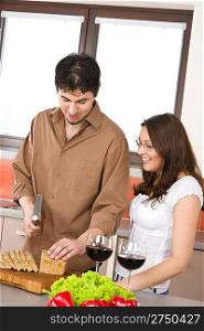 Happy couple cut bread in modern kitchen together and drink red wine