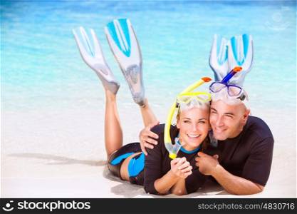Happy couple enjoys beach activities, tourists lying down on sandy coast and hugging each other, wearing snorkeling gear, water sport, spending honeymoon vacation on beach resort