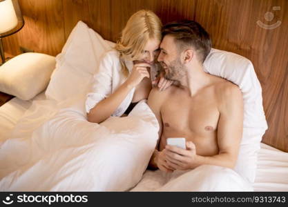 Happy couple in bed at home and use mobile phone