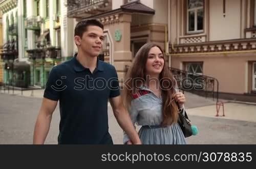 Happy couple in love on a date walking through city street holding hands. Handsome hipster man and attractive girlfriend holding hands strolling along old city street, enjoying romantic date and smiling. Slow motion. Steadicam stabilized shot.