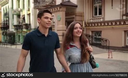 Happy couple in love on a date walking through city street holding hands. Handsome hipster man and attractive girlfriend holding hands strolling along old city street, enjoying romantic date and smiling. Slow motion. Steadicam stabilized shot.