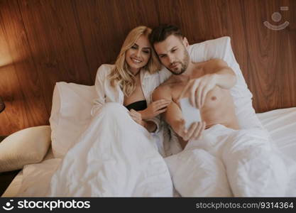 Happy couple in lying in bed at home and use mobile phone
