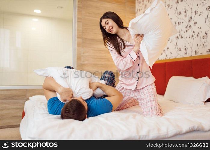 Happy couple in pajamas fights with pillows in bedroom at home, good morning. Harmonious relationship in young family. Man and woman resting together in their house, carefree weekend. Happy couple in pajamas fights with pillows