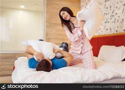 Happy couple in pajamas fights with pillows in bedroom at home, good morning. Harmonious relationship in young family. Man and woman resting together in their house, carefree weekend. Happy couple in pajamas fights with pillows