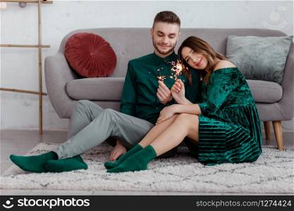 happy couple in stylish green clothes with sparklers. Man and pregnant woman sitting near sofa. Valentine&rsquo;s Day.. happy couple in stylish green clothes with sparklers. Man and pregnant woman sitting near sofa. Valentine&rsquo;s Day