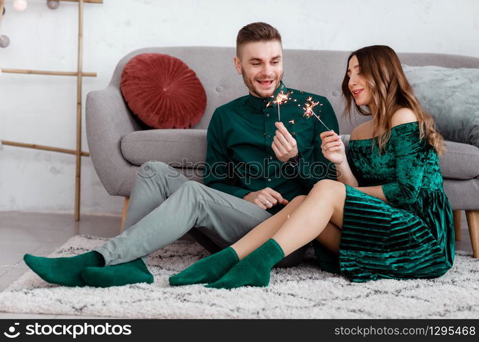 happy couple in stylish green clothes with sparklers. Man and pregnant woman sitting near sofa. Valentine&rsquo;s Day.. happy couple in stylish green clothes with sparklers. Man and pregnant woman sitting near sofa. Valentine&rsquo;s Day