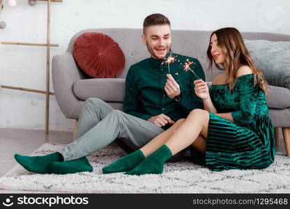 happy couple in stylish green clothes with sparklers. Man and pregnant woman sitting near sofa. Valentine&rsquo;s Day.. happy couple in stylish green clothes with sparklers. Man and pregnant woman sitting near sofa. Valentine&rsquo;s Day