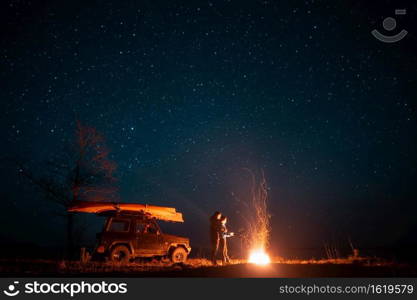Happy couple man and woman standing in front burning bonfire under starry sky, enjoying quiet night. Happy couple man and woman standing in front burning bonfire