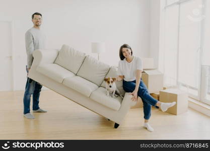 Happy couple move furniture in their new modern home, carry sofa with pet, pose in spacious room, lift couch in living room, have happy looks, celebrate moving day, carton boxes on floor near