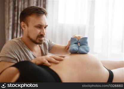 Happy couple, pregnant wife and husband plays with clothing for newborns on belly at home, bedroom interior on background. Pregnancy, prenatal period. Expectant mom and dad are resting in bed. Pregnant couple plays with clothing for newborns