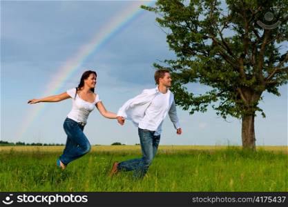 Happy couple running on a meadow in summer; in the background a rainbow