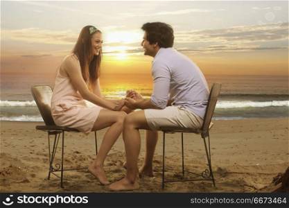 Happy couple sitting in chairs holding hands on beach at sunset