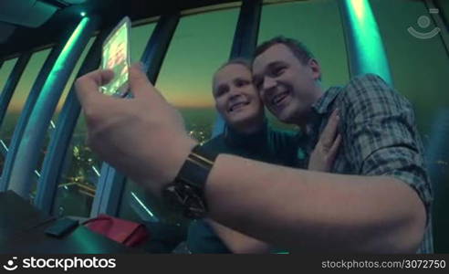 Happy couple taking selfie for smartphone being in night club or restaurant. Overlooking view of the night city is on the background.