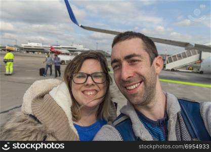 Happy couple taking selfie with smartphone or camera in airport outside near airplane.