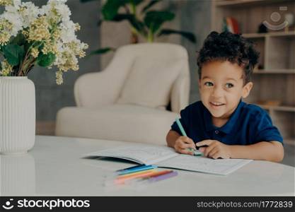 Happy curly mixed race boy coloring animals in coloring book with felt pens, looking aside and smiling while sitting in front of table in comfy living room at home. Children leisure time activities. Cute afro american child painting in coloring book at home