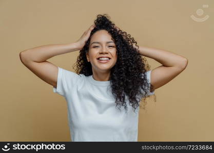 Happy curly woman keeps hands on head, has eyes full of happiness, smiles pleasantly and enjoys life, dressed in snow white t shirt, poses against beige studio background, glad to hear awesome news