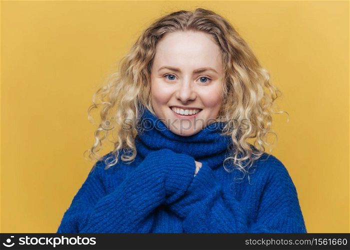 Happy curly young woman with broad smile, expresses positive emotions, wears blue sweater with collar, poses against yellow background. Good looking female model satisfied to hear good news.