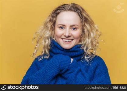 Happy curly young woman with broad smile, expresses positive emotions, wears blue sweater with collar, poses against yellow background. Good looking female model satisfied to hear good news.