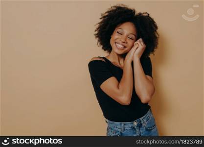 Happy cute 20s african woman with curly hair keeping hands near face and eyes closed, smiling pleasantly while dreaming of something good, isolated over beige background. Human emotions concept. Cute woman keeping hands near face and dreaming of something good, isolated over beige wall