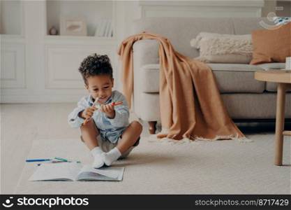 Happy cute african boy holding two felt tip pen, choosing which color to use for drawing in his new coloring book, kid being very excited while spending leisure time and playing in living room at home. Cute afro american boy kid with colouring book sitting on floor in cozy living room