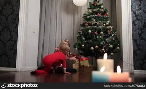 Happy cute girl and her toddler brother in santa costume opening Xmas presents. Children under Christmas tree with gift boxes. Christmas decorated living room with traditional candles and decorations. Cozy warm winter evening at home.