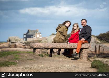happy dad and two daughters sits on a bench near the sea and the shore at the Tregastel, Brittany. France