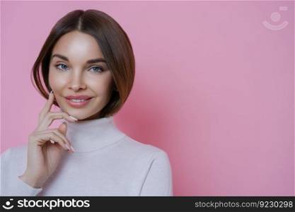 Happy dark haired woman with well groomed complexion, smiles tenderly, touches cheek, has manicure, pretty face, dressed in white turtleneck, isolated on pink background. Natural beauty concept