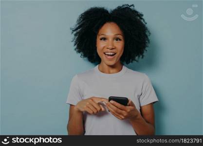 Happy dark skinned girl holding modern smartphone and chatting with friend online or using mobile application, pointing at device screen and looking at camera with excited face expression. Beautiful happy dark skinned girl holding modern smartphone and chatting with friend online
