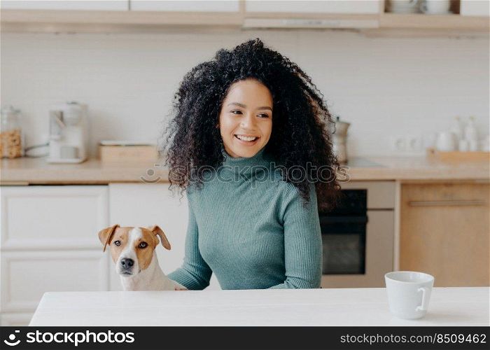 Happy dark skinned girl looks thoughtfully away, has pleasant smile, dressed in casual wear, drinks hot beverage, poses with domestic animal against kitchen interior. Female dog owner at home