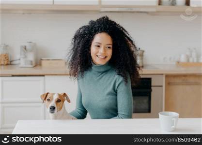 Happy dark skinned girl looks thoughtfully away, has pleasant smile, dressed in casual wear, drinks hot beverage, poses with domestic animal against kitchen interior. Female dog owner at home